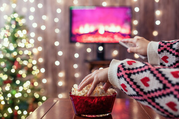 Christmas eve. Alone woman eating popcorn, watching tv, switching channels using remote control. Home cinema. Cropped, close up