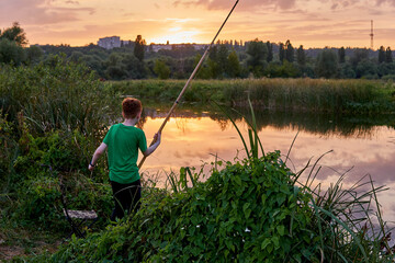 Young boy fishing with the pole near the lake on the sunset