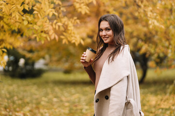 A beautiful pretty young woman in a coat drinks hot coffee walks in the autumn park smiles communicates online and talks using a mobile phone in nature in the fall forest, selective focus