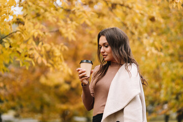 A beautiful pretty young woman in a coat drinks hot coffee walks in the autumn park smiles communicates online and talks using a mobile phone in nature in the fall forest, selective focus