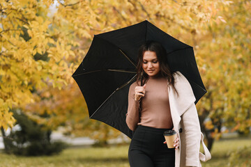 A pretty happy young woman in a casual coat enjoys solitude walking holding a black umbrella in rainy weather in an fall park in nature in autumn, selective focus