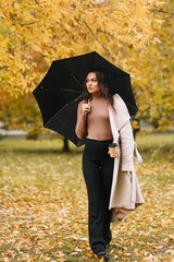 A pretty happy young woman in a casual coat enjoys solitude walking holding a black umbrella in rainy weather in an fall park in nature in autumn, selective focus