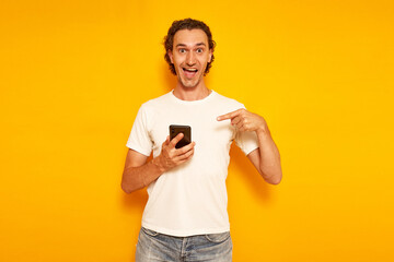an excited happy man in casual clothes with phone in his hand points his finger at smartphone and we are happy to receive message about discounts, promotions and sales. isolated on yellow background.