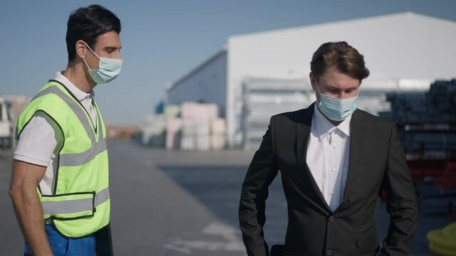 Medium shot of Middle Eastern and Caucasian men in coronavirus face masks touching elbows walking away in slow motion. Warehouse manager in uniform and supervisor inspector in suit outdoors