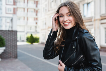 happy young woman in black turtleneck and leather jacket adjusting hair on urban street of europe