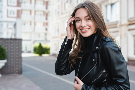 happy young woman in black turtleneck and leather jacket adjusting hair on urban street of europe - Powered by Adobe