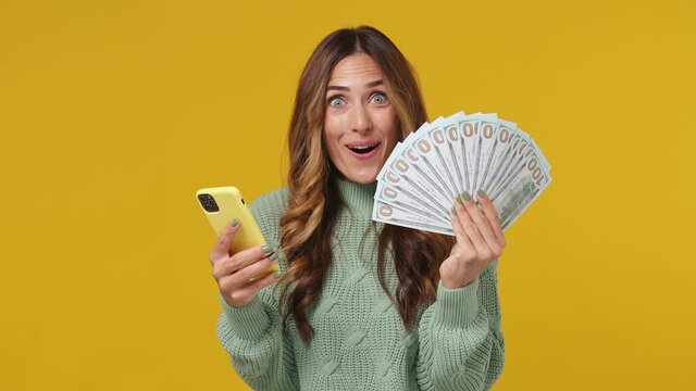 Excited overjoyed joyful young brunette woman 30s years old wears mint sweater using mobile cell phone hold fan of cash money in dollar banknotes isolated on plain yellow background studio portrait