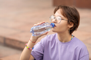 a brunette teenage girl with glasses on the streets of the city drinks water from a bottle on a clear sunny day