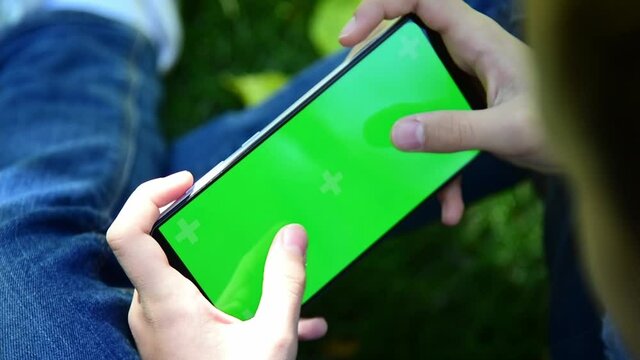 The child is holding a phone with a green screen in nature. A girl lies in the outumn on the grass in a blue dress and looks at the phone