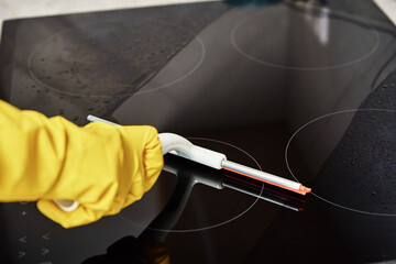 Cleaning induction stove. Woman in yellow rubber gloves cleans kitchen induction hob with cleaning...