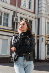 dreamy young woman in leather jacket standing on urban street