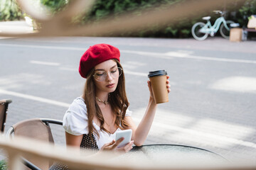 young woman in red beret and eyeglasses holding paper cup and using smartphone on terrace of cafe
