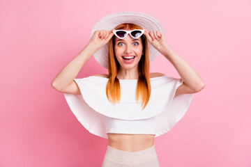 Portrait of attractive amazed cheerful red-haired girl touching specs resort isolated over pink pastel color background