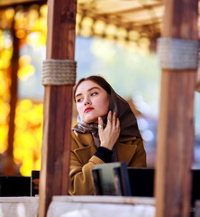 Luxurious young woman in retro fashion style, in beige coat and scarf on her head, in a cafe on wooden playground in autumn