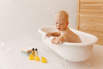 Bathing the baby in white bath with yellow rubber ducks, towels. Baby sitting in the bath and looking at the camera. A lot of bubbles flying in the bathroom.