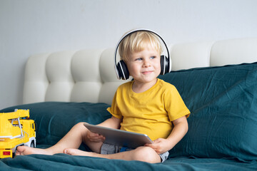 Smiling happy little kid boy using tablet in wireless headphones in bed at home