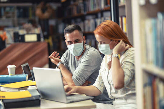 Couple of friends studying in campus library. They're wearing face masks during pandemic restrictions.	