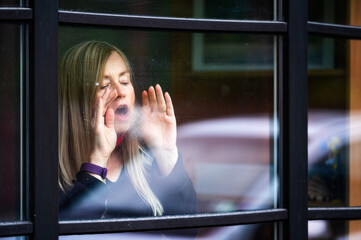 closeup portrait of middle aged blonde woman staying behind window, breathing on the glass