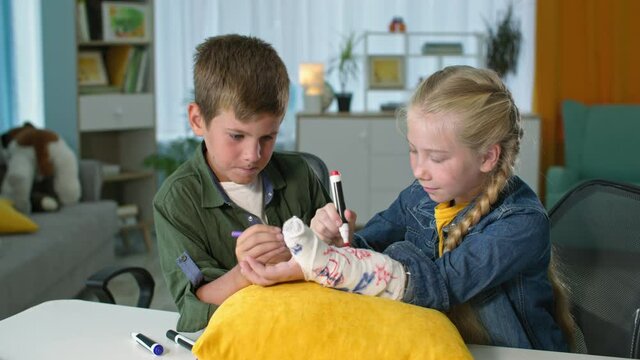 child cares and supports friend, kids with multicolored felt-tip pens paint plaster cast on arm, boy and girl show thumbs up at home