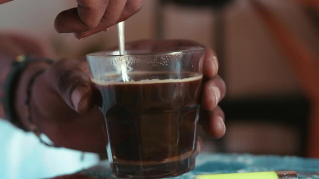 Photo of a person stirring sugar with a metal spoon in a glass of coffee