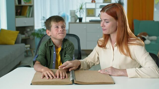 blind boy learning to read braille book while sitting next to a female teacher at home, fingers of kid move on a sheet of paper
