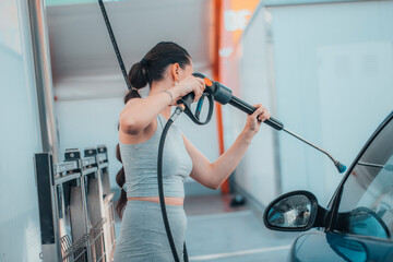 Chica joven trabajadora en un lavadero de coches limpiando con una manguera de agua a presión el vehículo de un cliente