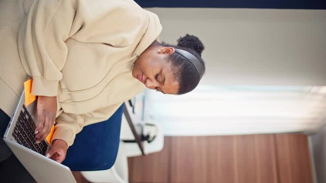 Vertical Portrait Of Plump Black Woman In Beige Hoodie Typing On Laptop Pc, Looking At Camera And Smiling While Working From Home. Female Working Or Studying Remotely During Self-isolation Quarantine