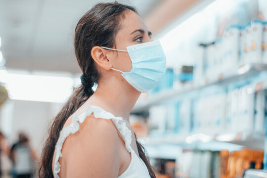 Mujer joven con mascarilla quir&uacute;rgica observando interesada en las estanter&iacute;as de la farmac&eacute;utica para comprar un f&aacute;rmaco de medicinas en el comercio