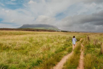  Young slim teenager girl walking on a small road towards Strandhill town, Knocknarea hill in cloudy sky in the background. County Sligo, Ireland. Irish landscape. Popular landmark. © mark_gusev