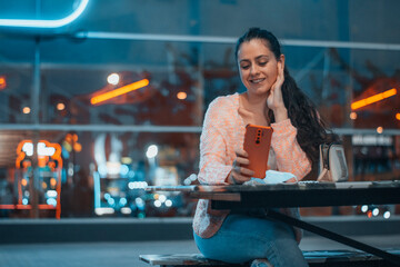 Adolescente sonriente feliz haciéndose fotografías junto a las cristaleras del centro de ocio mientras espera sentada en la mesa esperando ser atendida para cenar con su acompañante