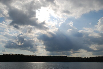 Formidable cumulus clouds over the lake. Small waves on the surface of the water. Above the water and the forest standing on the other side, white-gray clouds, behind which the sun is partially hidden