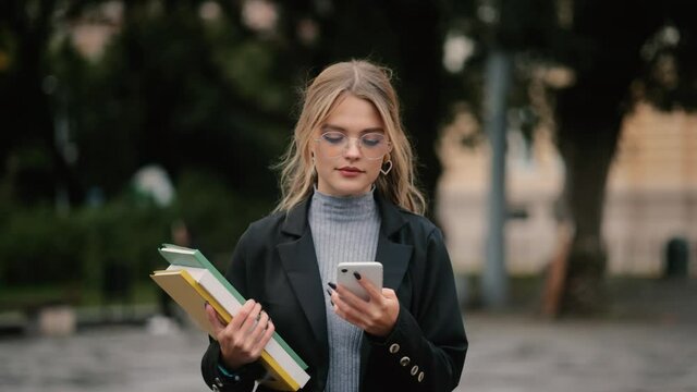 Student girl in glasses with books walking to the university building. Portrait of attractive girl texting on her phone. High quality 4k footage