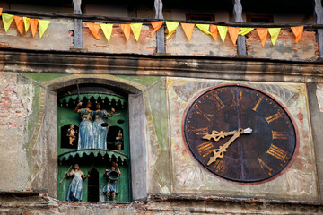 Sighisoara clock tower, left with medieval puppet play, clock with rusty clock face, yellow-orange roman numerals and metal clock hands right side and yellow orange pennant chain above