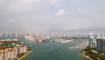 Fototapeta premium Incredible aerial view over the Miami shipping channels with the skyline on the horizon beyond and cloudy sky above as boats and ferries make their way to and from Fisher Island below.
