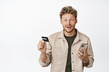 Handsome blond curly guy holding smartphone and credit card, looking excited about store sale, paying online, order in mobile phone, white background