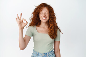 Fototapeta na wymiar Positive beautiful girl with ginger hair, winking and smiling pleased, shows okay, ok sign, recommending something, like and approve, white background
