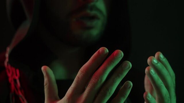 Evil exorcist reading spell close-up. Wicked shaman magician wearing robe and chains standing in darkness, making ritual, scary wizard using witchcraft, black magic. Halloween concept. 