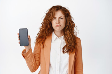 Naklejka premium Sad frowning business woman showing mobile phone screen and sulking, dislike smartphone app, standing in suit against white background