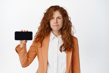 Skeptical business woman shows horizontal phone screen and sulking disappointed, dislike...