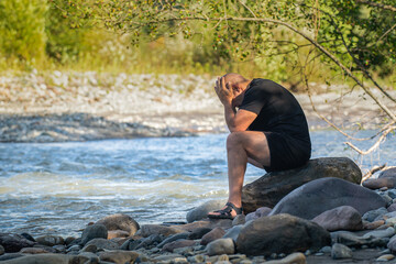 A young man, upset with something, sits on the bank of a mountain river, his head clasped in his hands.