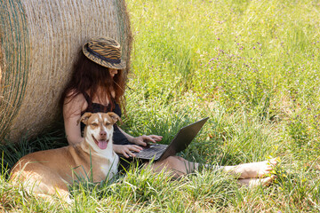 woman with red hair near a haystack works with a laptop next to her her dog