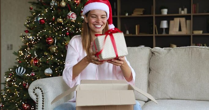 Receiving New Year gifts. Excited young woman in funny santa hat sit on sofa near Christmas tree unbox parcel take out two wrapped presents. Happy gen z lady get cute Xmas surprise in postal package