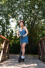 A girl in a safety helmet rides an electric scooter across a wooden bridge. There is beautiful nature around. Summer sunny day. Leisure activities, sports, environmentally friendly transport.