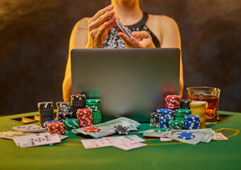 A young elegant woman sits at a gambling table on which cards and stacks of chips are laid out. Open laptop. Concept - online casino, gambling, poker game, game strategy.