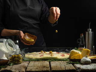 The chef lays out the olives on the glass dough prepared for baking. Ingredients. Cooking Italian focaccia bread, pizza. Wooden texture, black background. Close-up. Restaurant, hotel, pizzeria.