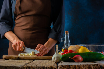 The chef cuts the cheese on a cutting board. Ingredients for making a classic Greek salad. Close-up. Wooden texture. Vitamin dietary dish. Low in calories. Restaurant, hotel, home cooking.