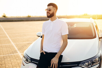 Fototapeta na wymiar Young bearded man standing near a car outdoors on a summer day, a man in a white shirt stands next to a white car