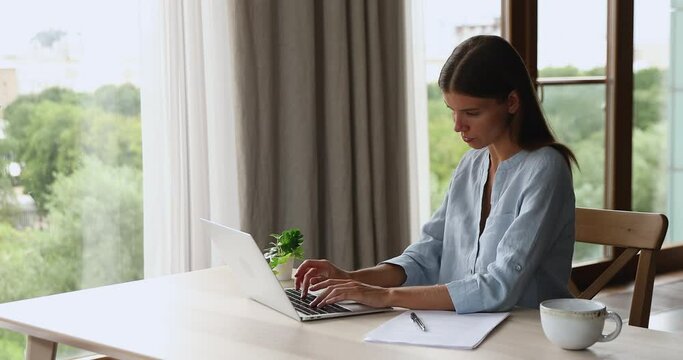 Focused young woman sit by home office desk search compare data online using laptop. Inspired female designer work on pc in graphic editor app develop new project model combine patterns on screen