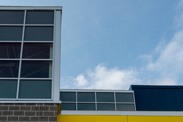 Exterior corner of a building with glass windows, a wall of yellow metal composite panels, a dark brown brick wall base with grey mortar. The background is blue sky with white clouds on a sunny day.