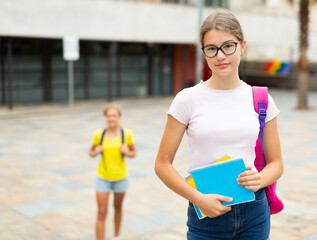 Young girl with copybooks in hand standing near school building outdoors. Her classmate walking behind.